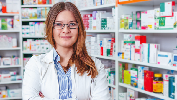 pharmacy technician in a lab coat in front of a shelf of prescriptions