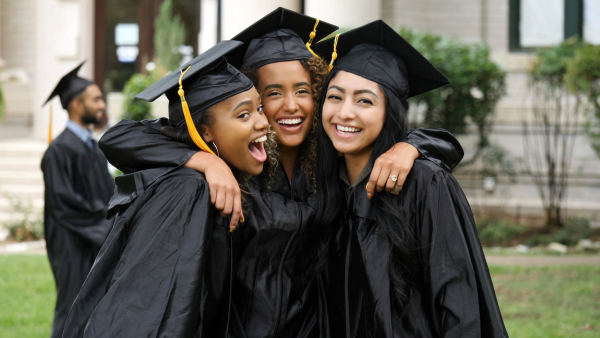 Three women standing together and smiling at each other in graduation attire. 
